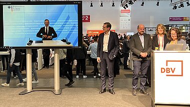 Bildschirm einer Liveübertragung und Podium mit Redenden