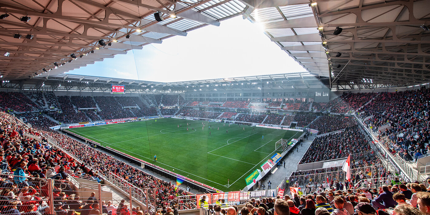 Stadion Innenansicht mit gefüllten Zuschauerplätzen im Vordergrund und Spielfeld im Hintergrund